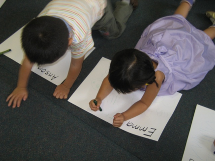 Two preschoolers lying on their stomachs, practising writing thier names on large paper.