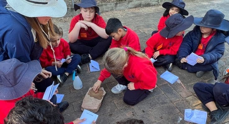 A group of students sitting in a circle outside, watching one student making paint from ochre.