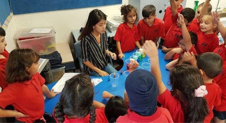 Teacher performing a science experiment at a curved table with students watching. Some students have hands up to answer questions.