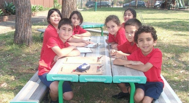Seven students sitting at an outdoor picnic table doing school work.